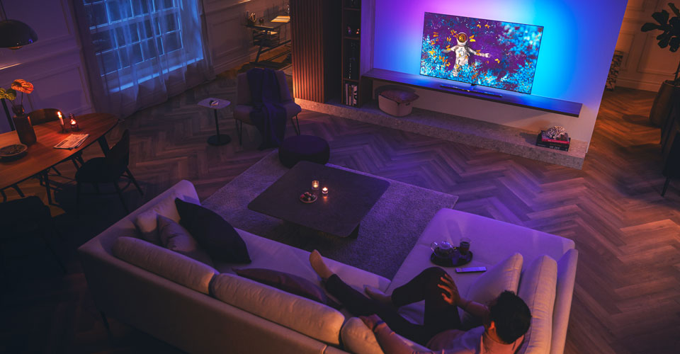 Mees l&otilde;&otilde;gastub h&auml;maralt valgustatud elutoas diivanil ja vaatab Philipsi Ambilight-telerit